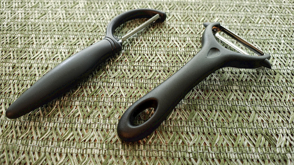 Seitliche Ansicht der beiden Yoosso-Schäler auf strukturierter Oberfläche, die die ergonomischen Griffe und die unterschiedlichen Klingenformen deutlich erkennbar macht.